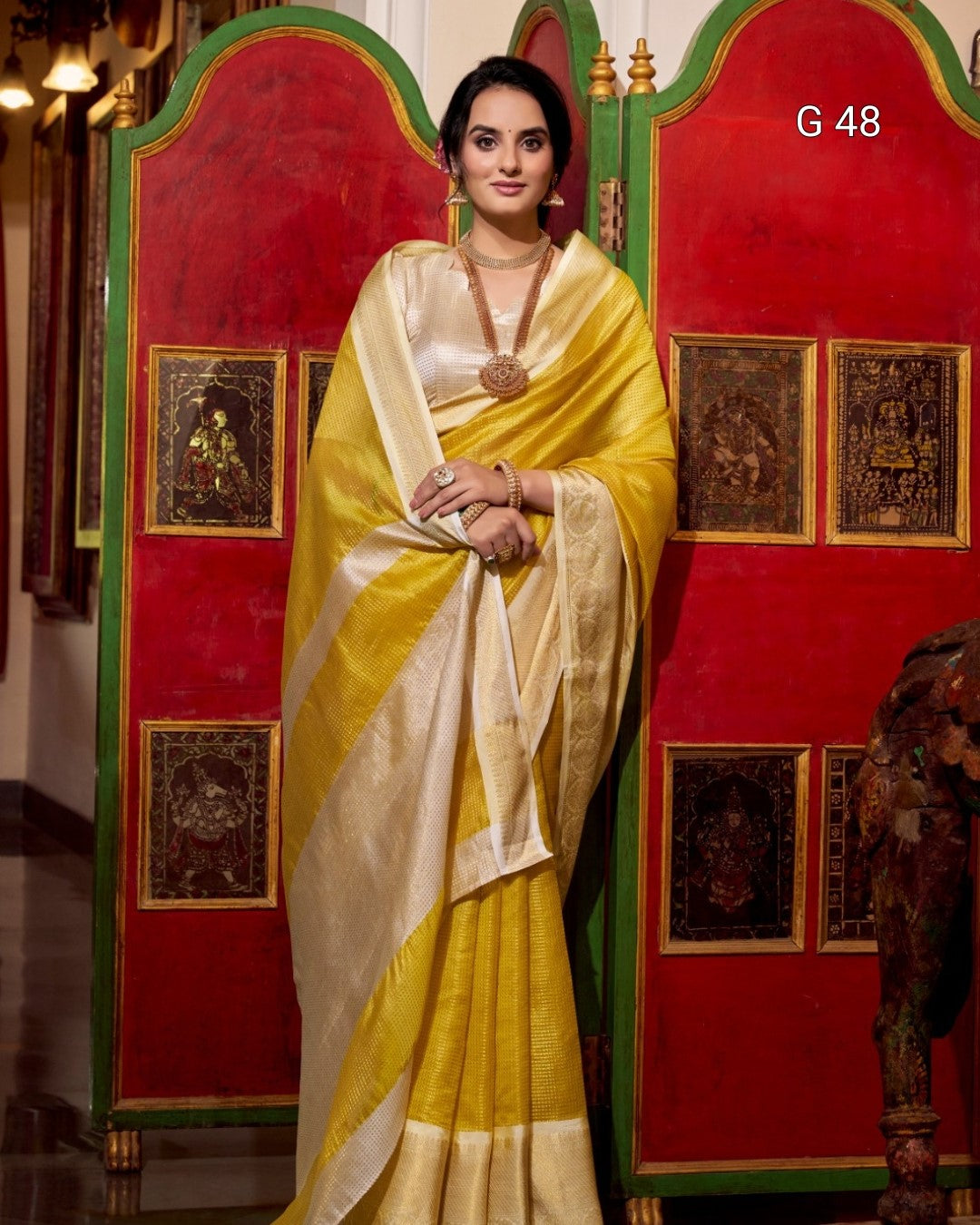 Woman in a yellow saree standing in front of a decorative red and gold screen.