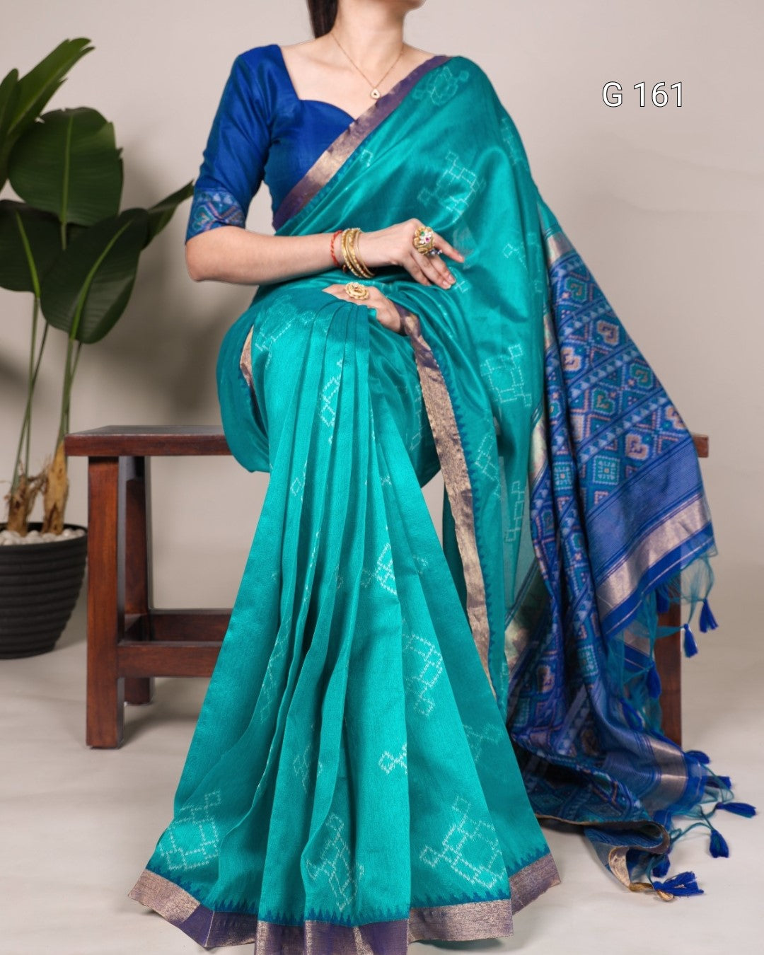 Teal saree with blue blouse draped over a person, with a plant and wooden stool in the background.