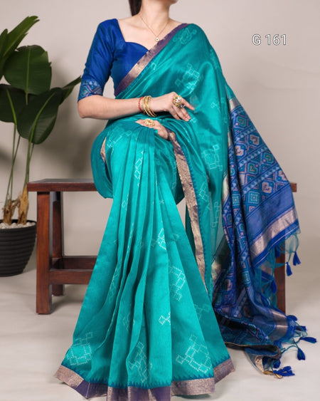 Teal saree with blue blouse draped over a person, with a plant and wooden stool in the background.
