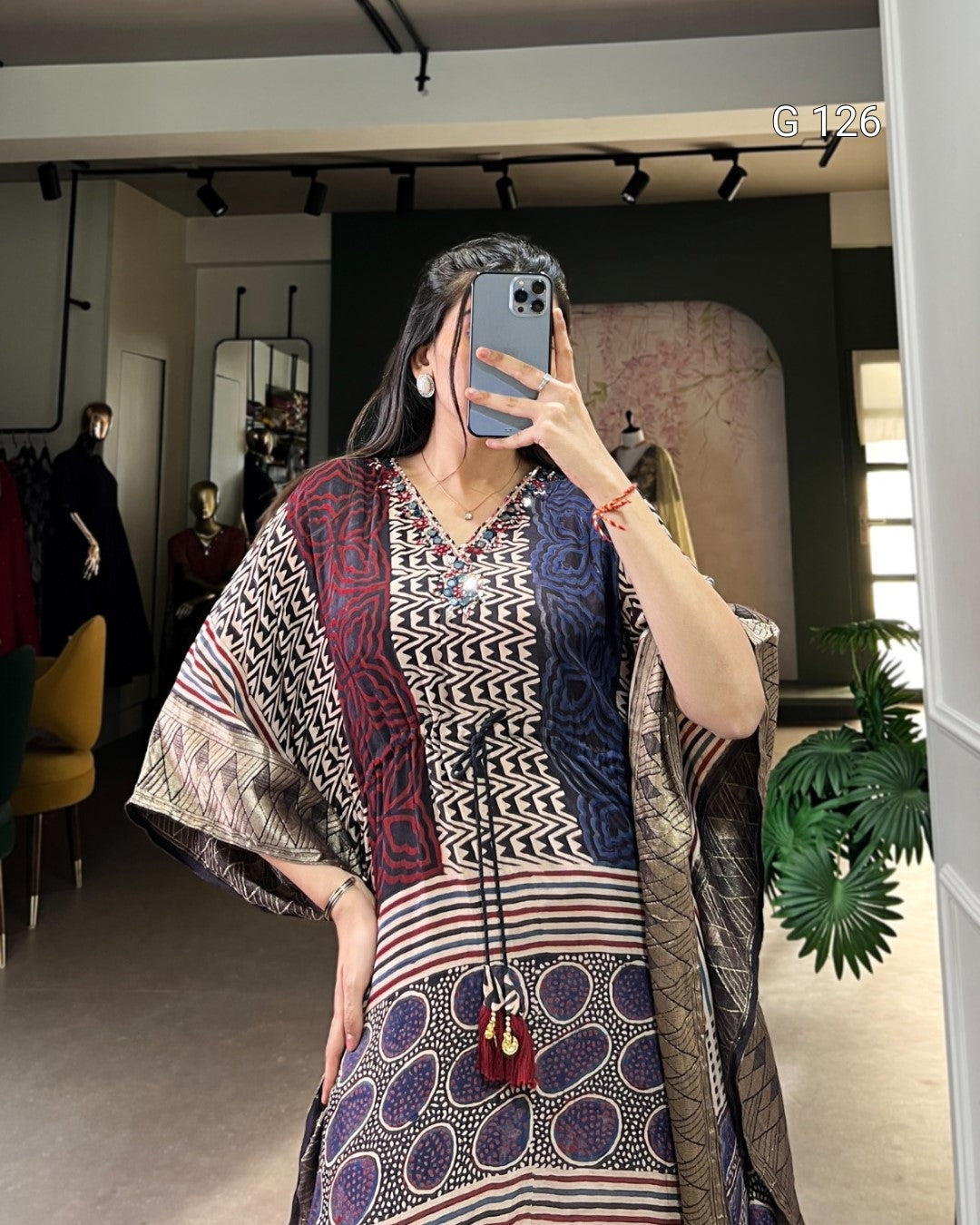 Person wearing a patterned outfit with a long scarf in a room with plants and furniture.