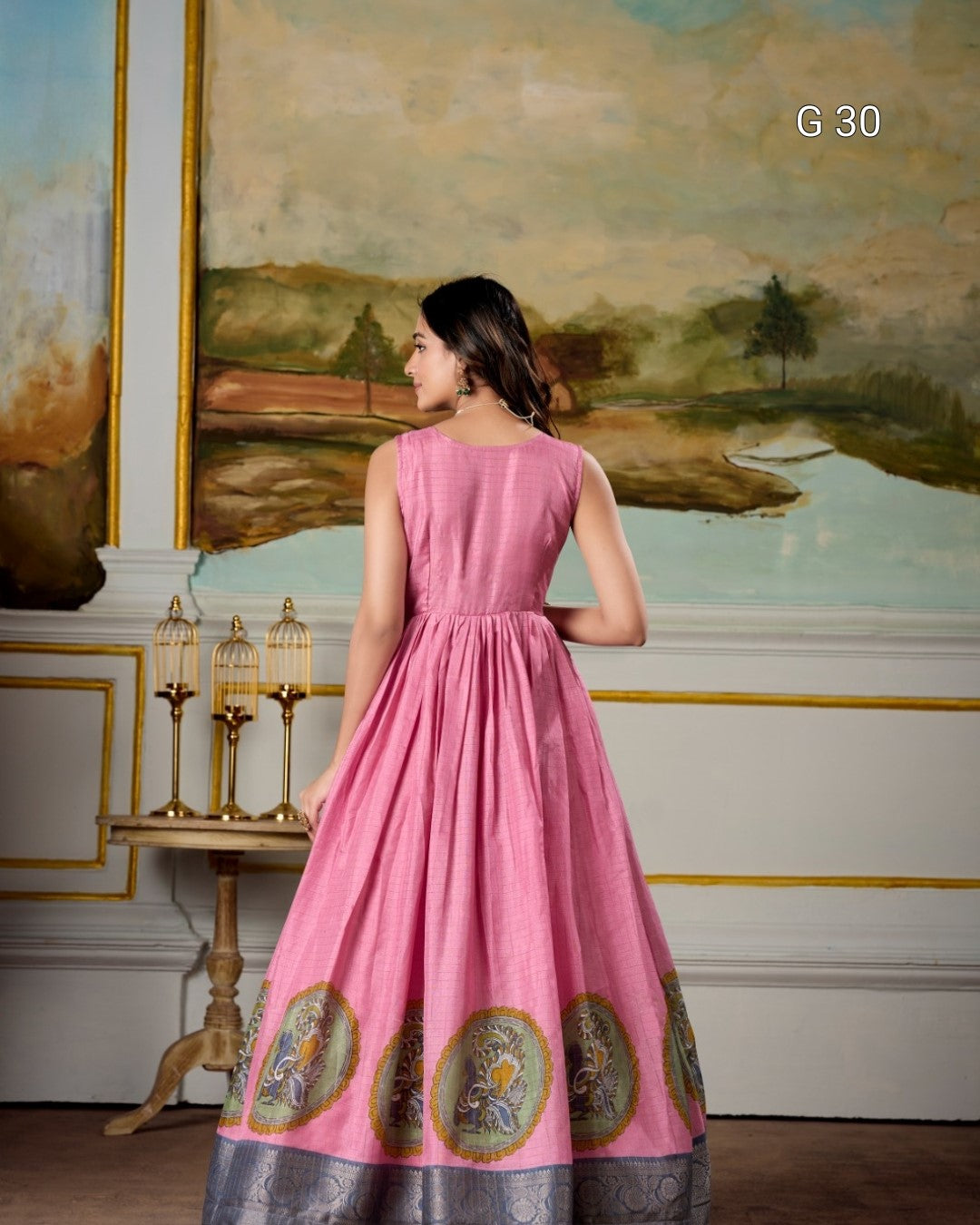 Woman in a pink dress with gold patterns standing in a room with a painting on the wall.