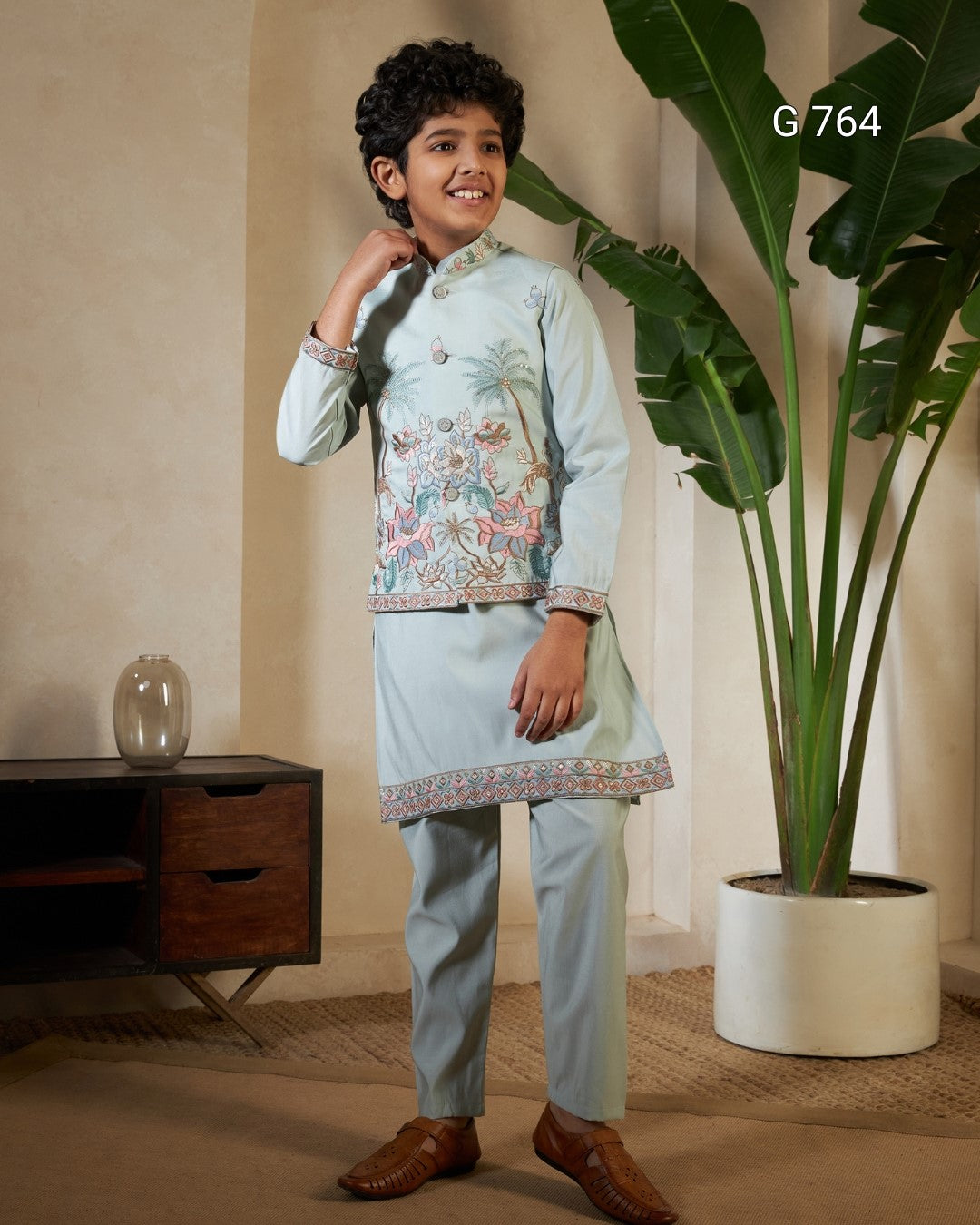 Child wearing a light blue embroidered traditional outfit standing indoors with a plant and wooden cabinet in the background.