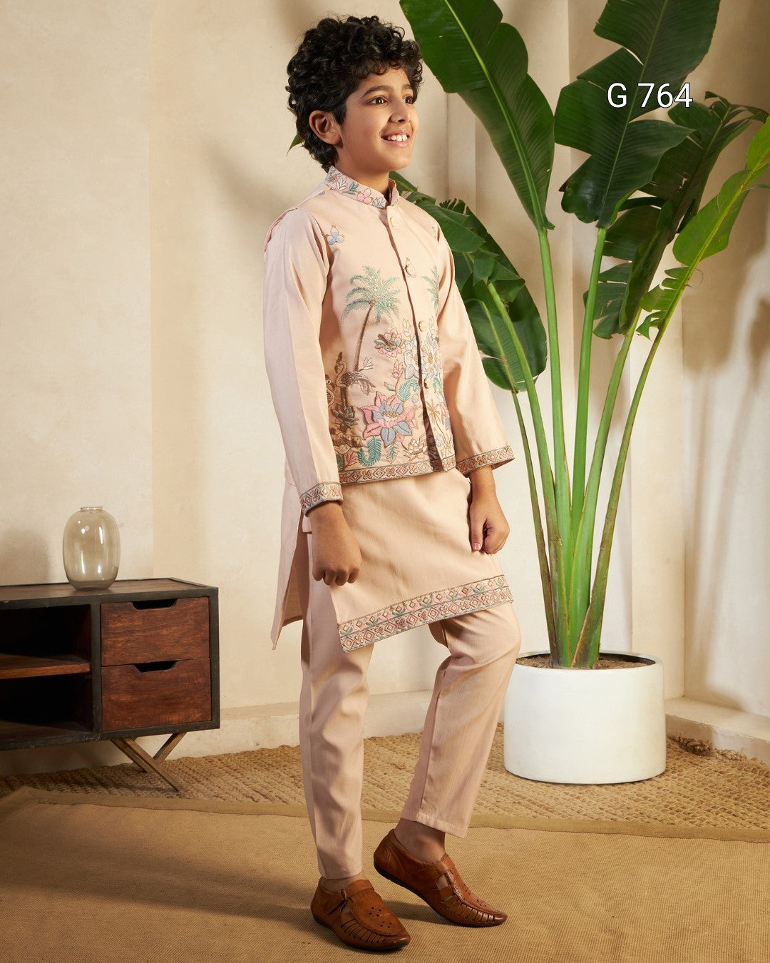 Young boy in traditional outfit standing in a room with a plant and wooden cabinet.