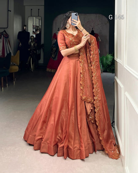 Woman in a traditional orange saree with intricate designs, standing in a room with mirrors and clothing in the background.