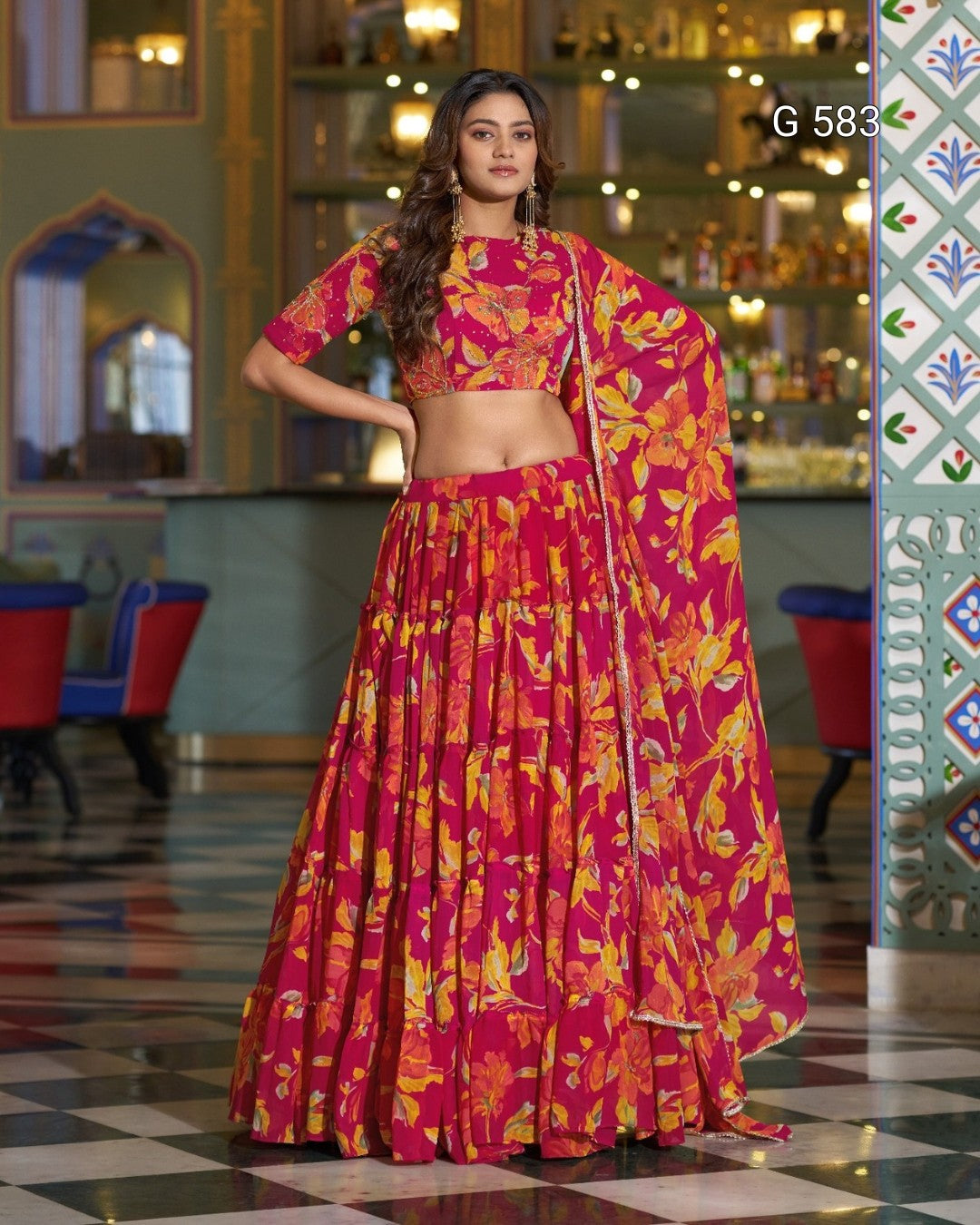 A woman models a traditional Indian lehenga choli set with a vibrant magenta, orange, and yellow floral print. The set includes a short-sleeved crop blouse, a matching flared skirt, and a draped dupatta.