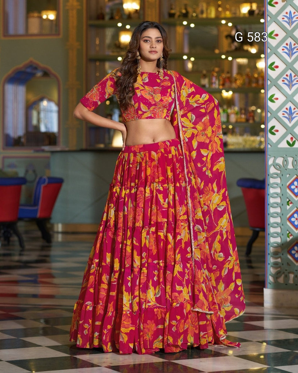 A woman models a traditional Indian lehenga choli set with a vibrant magenta, orange, and yellow floral print. The set includes a short-sleeved crop blouse, a matching flared skirt, and a draped dupatta.
