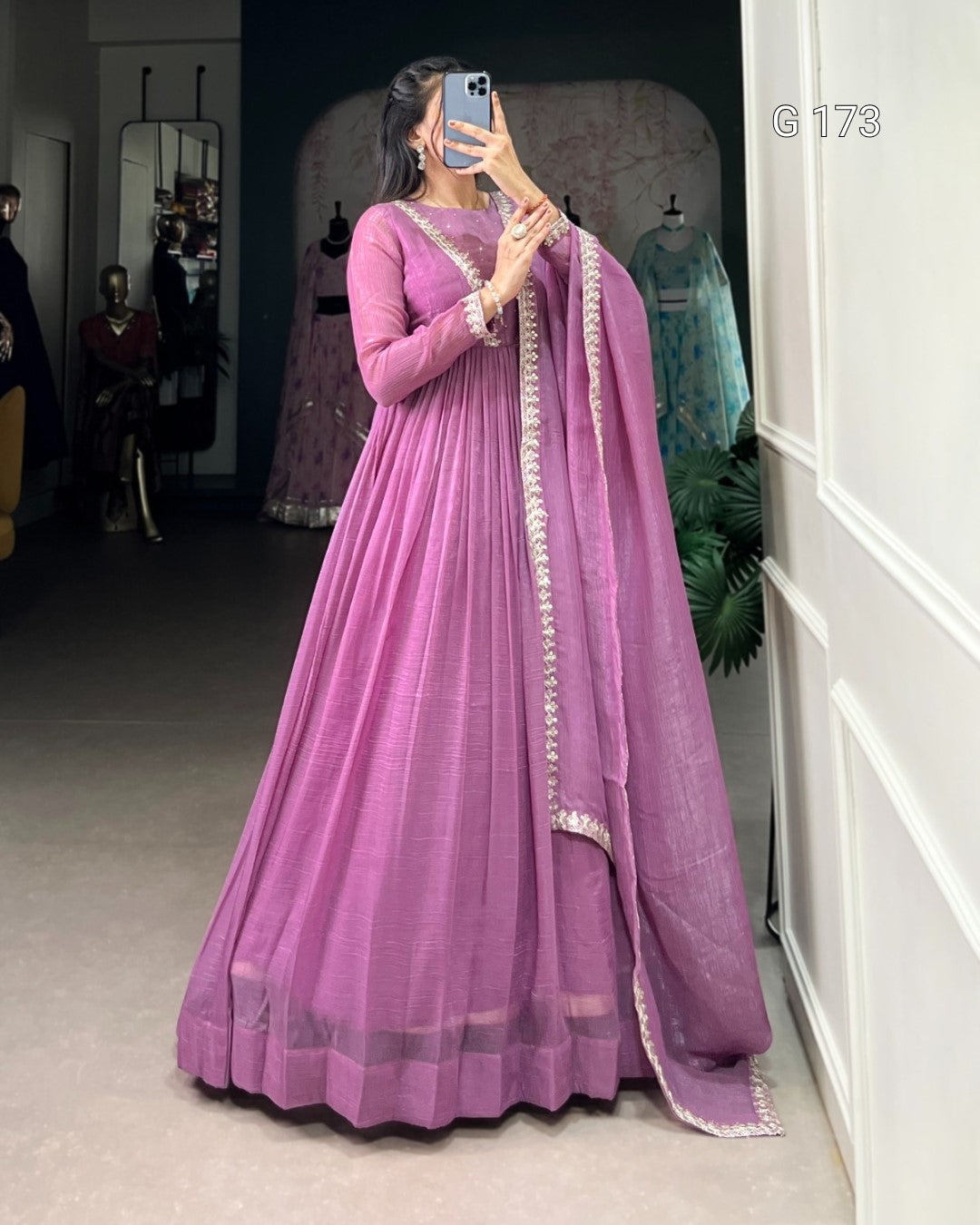 Woman wearing a purple traditional outfit with a matching dupatta, standing in a room with mirrors and clothing in the background.