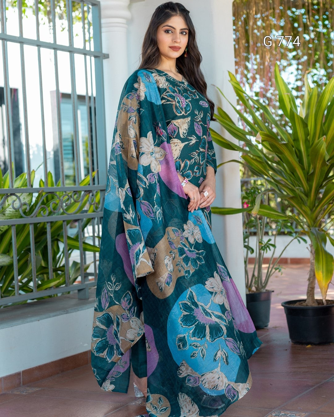 Woman wearing a teal floral saree with a background of plants and a railing.