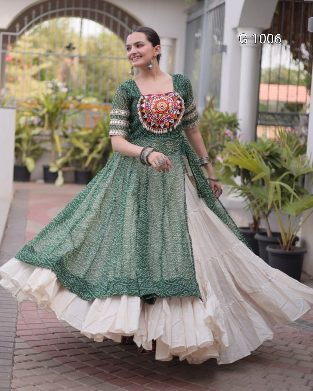 A woman wearing a traditional Indian outfit with a green embroidered kurta and a cream-colored flared skirt, standing in a garden courtyard.