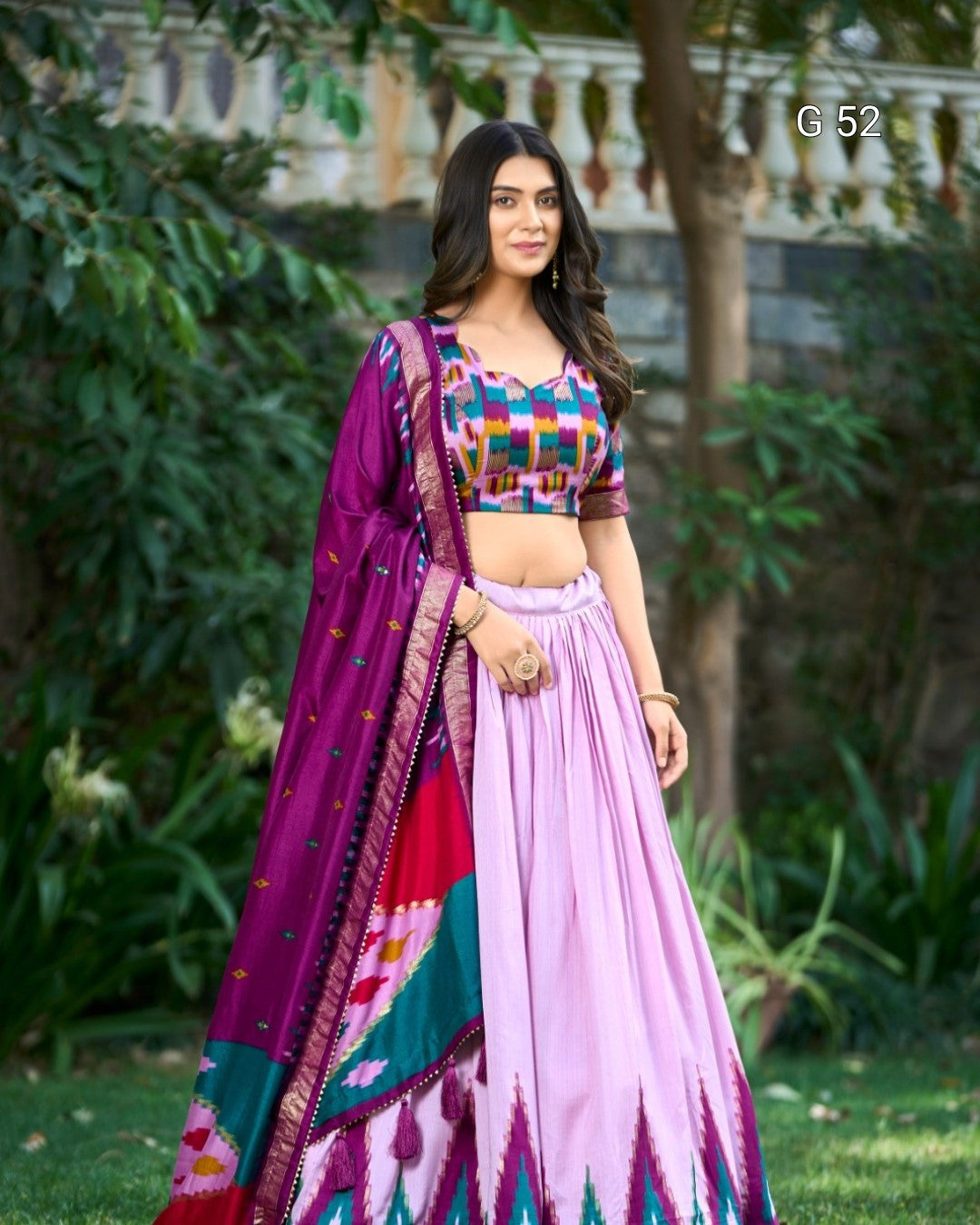 Woman in a colorful traditional outfit standing outdoors with greenery in the background