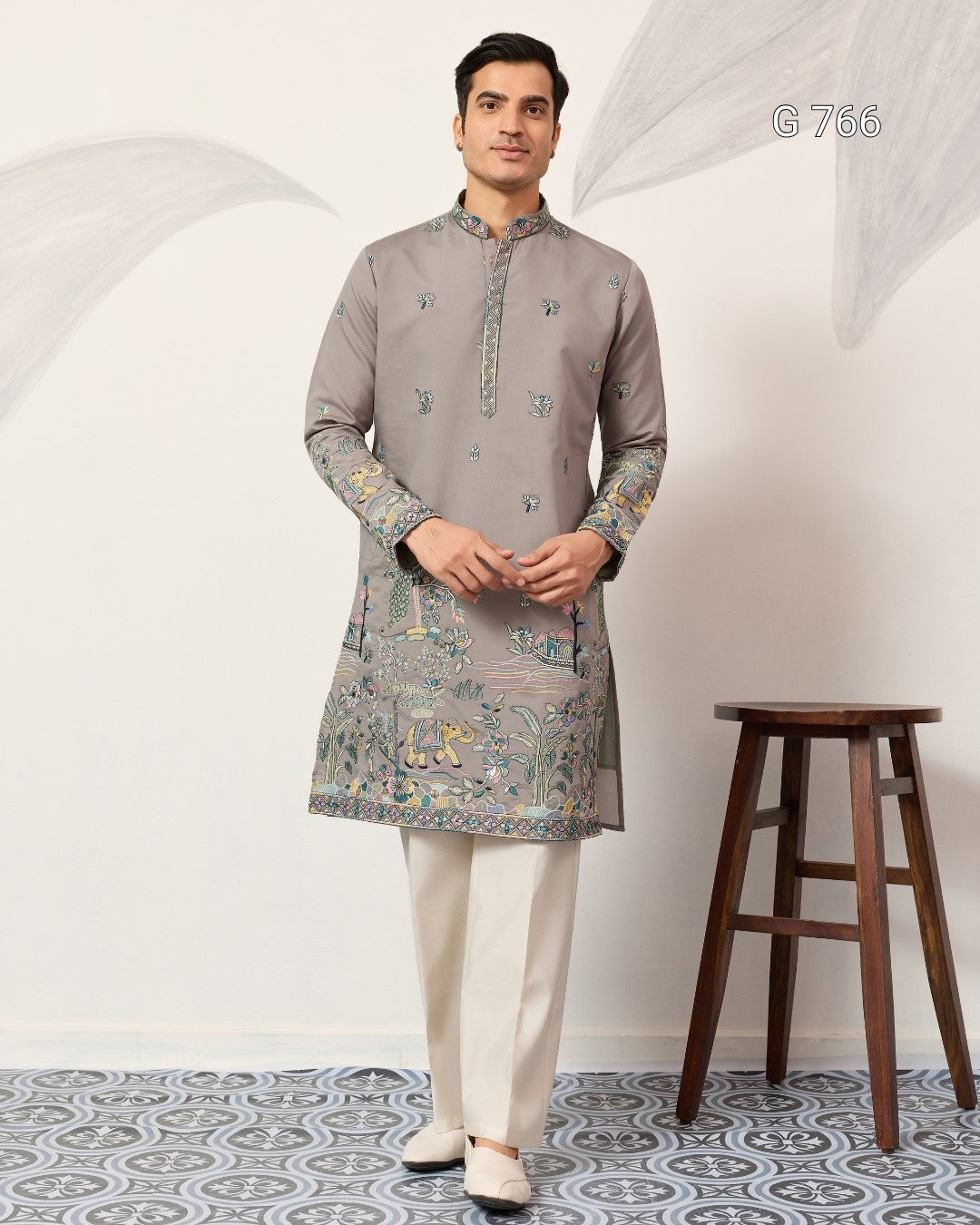 Man wearing a gray kurta with white pants standing in a room with a wooden stool.