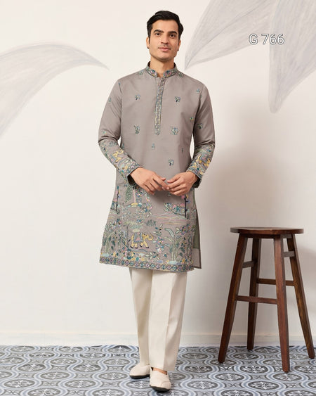 Man wearing a gray kurta with white pants standing in a room with a wooden stool.