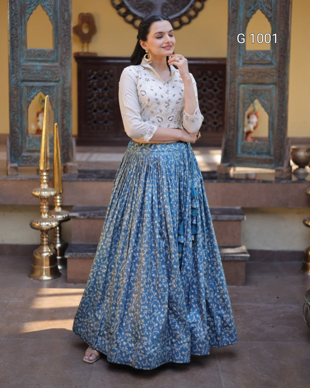 A woman modeling a traditional Indian outfit featuring a white embroidered kurta and a long blue floral printed skirt, standing in front of an ornate architectural background with yellow walls and carved wooden doors.