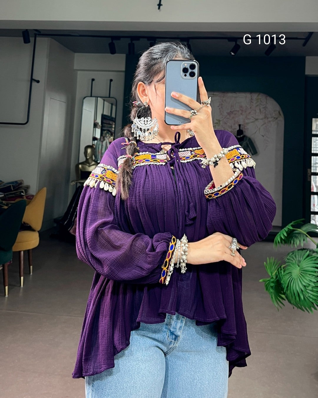 A woman takes a mirror selfie wearing a purple off-the-shoulder blouse with colorful embroidered details and silver-toned jewelry, paired with light blue jeans in an indoor boutique setting.