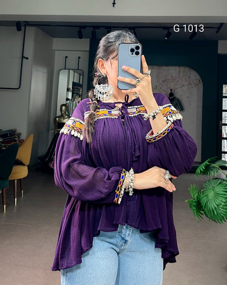 A woman takes a mirror selfie wearing a purple off-the-shoulder blouse with colorful embroidered details and silver-toned jewelry, paired with light blue jeans in an indoor boutique setting.