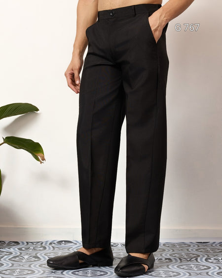 Person wearing black pants standing against a white wall with a plant on the left.