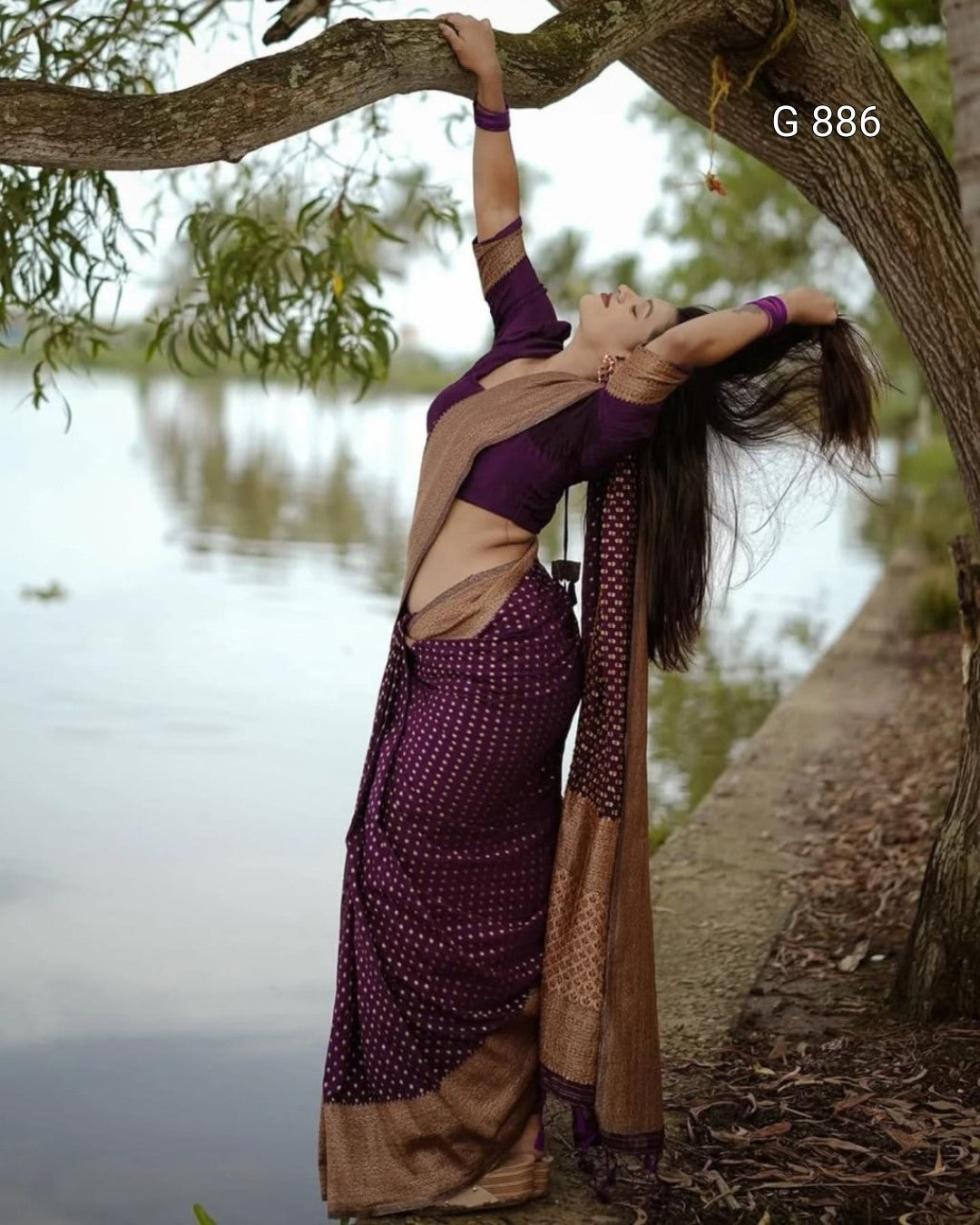 Woman in a purple and brown saree standing under a tree by a body of water.