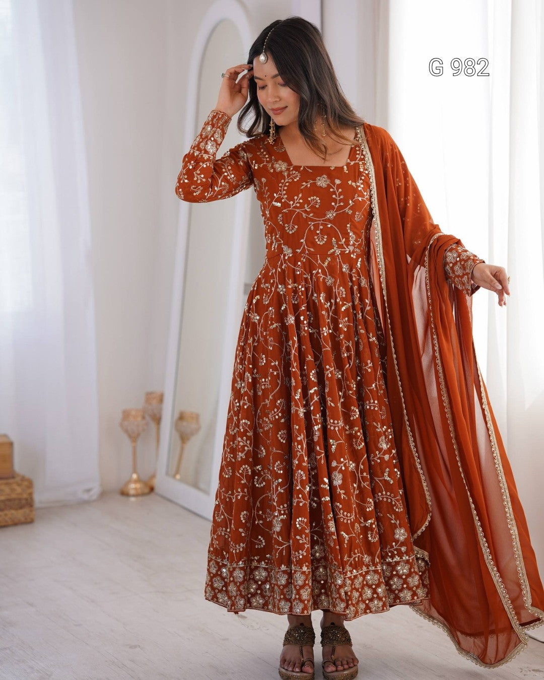 A woman models a rust-orange embroidered Anarkali gown with a matching sheer dupatta in a bright, white-walled room. The dress features intricate white floral embroidery and is styled with brown sandals.