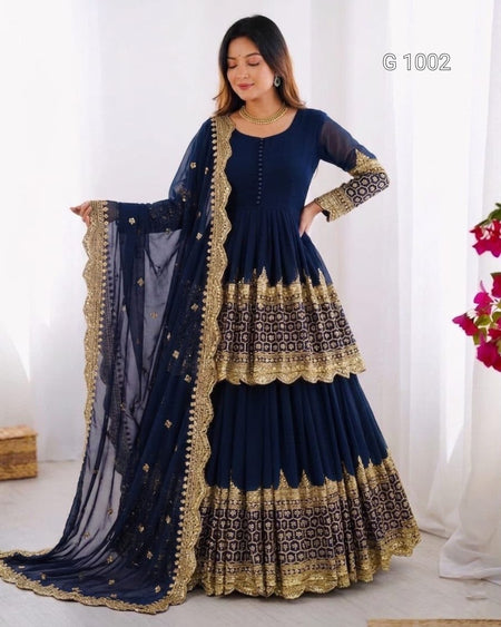 A woman models a navy blue traditional Anarkali gown with gold embroidery and a matching dupatta. The dress features a tiered skirt with intricate gold patterns and a scalloped border, sheer long sleeves, and a buttoned neckline.