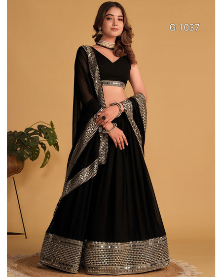 A woman models a black traditional lehenga choli set with ornate silver mirror work borders, set against a warm beige background.