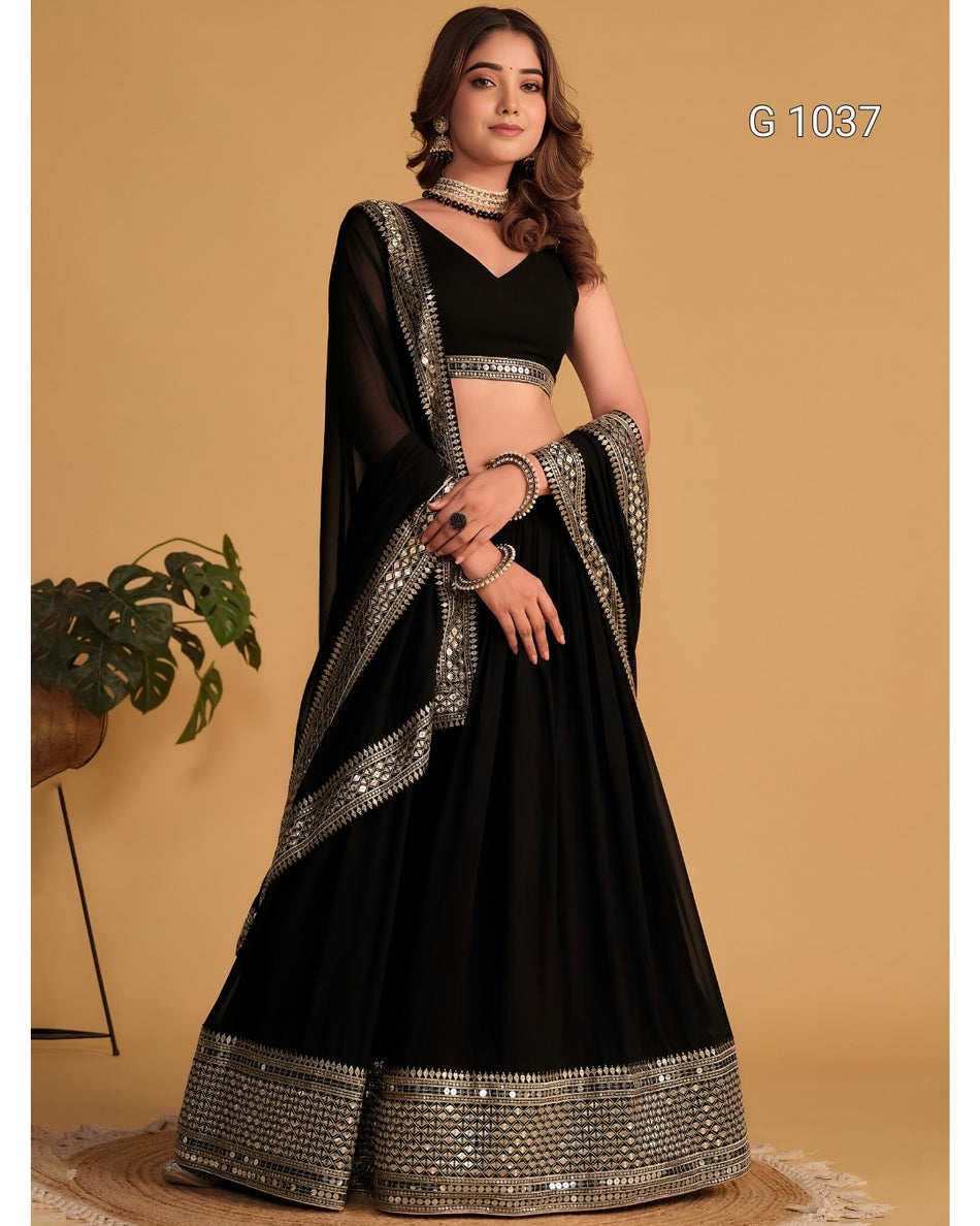 A woman models a black traditional lehenga choli set with ornate silver mirror work borders, set against a warm beige background.