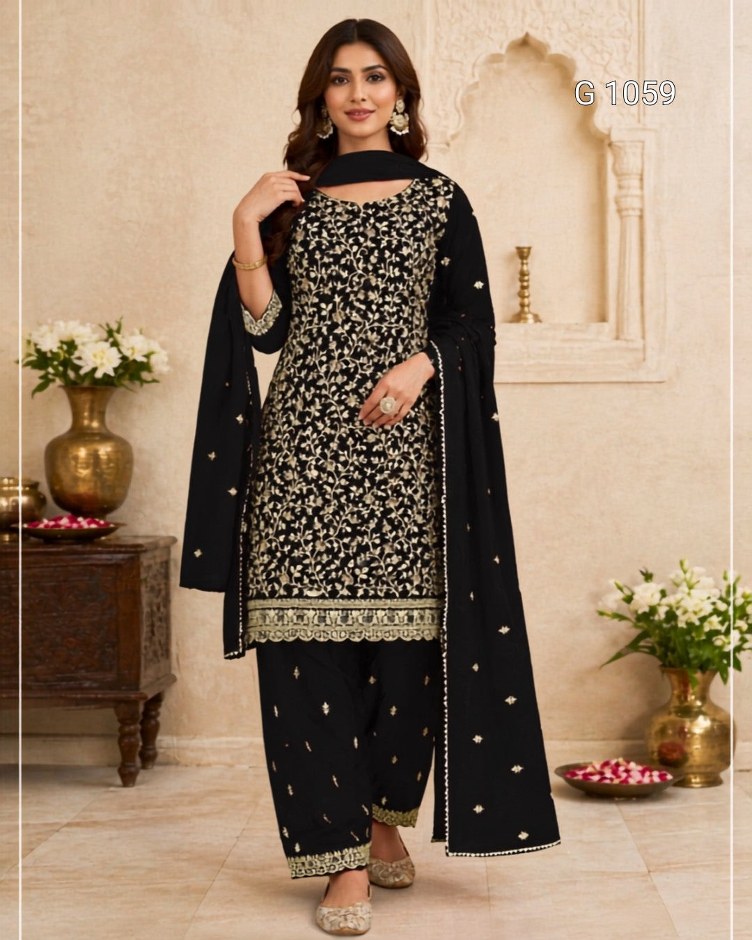 A woman models a black three-piece traditional outfit with intricate gold embroidery, featuring a kurta, pants, and dupatta, set against a warm beige architectural background with decorative pillars and flower arrangements.