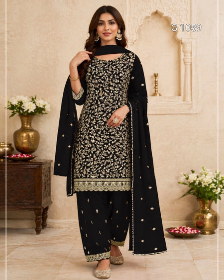 A woman models a black three-piece traditional outfit with intricate gold embroidery, featuring a kurta, pants, and dupatta, set against a warm beige architectural background with decorative pillars and flower arrangements.
