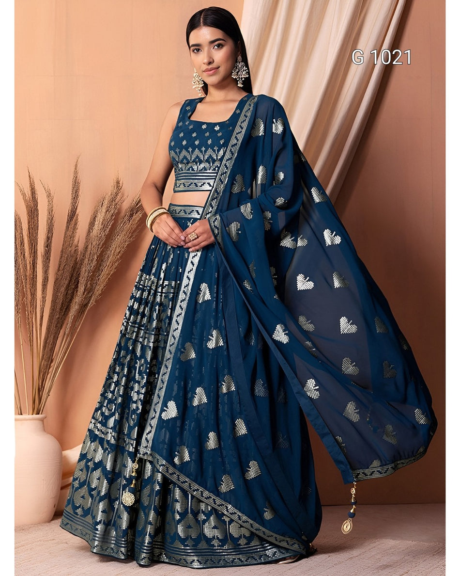 A woman models a dark blue traditional Indian outfit with metallic silver motifs and borders, featuring a sleeveless kurta, flared skirt, and matching dupatta, set against a warm beige background with decorative dried grass in a vase.