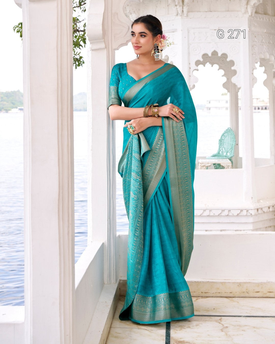 A woman models a teal blue-green silk saree with a metallic silver border, standing on a white marble balcony with water and a pavilion in the background.