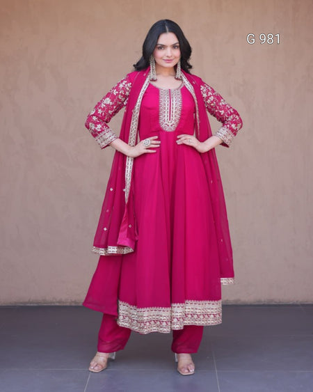 A woman modeling a magenta pink traditional Indian outfit, featuring a kurta with gold embroidery, matching pants, and a dupatta, set against a plain beige wall.
