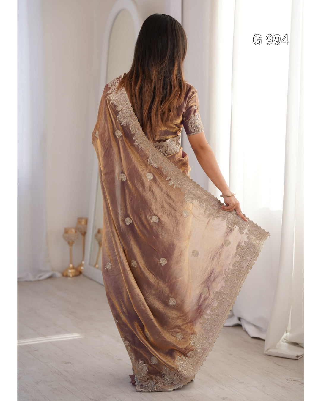 A woman viewed from behind wearing a metallic bronze-brown saree with a wide embroidered border and small floral motifs, standing in a bright indoor room with white curtains.