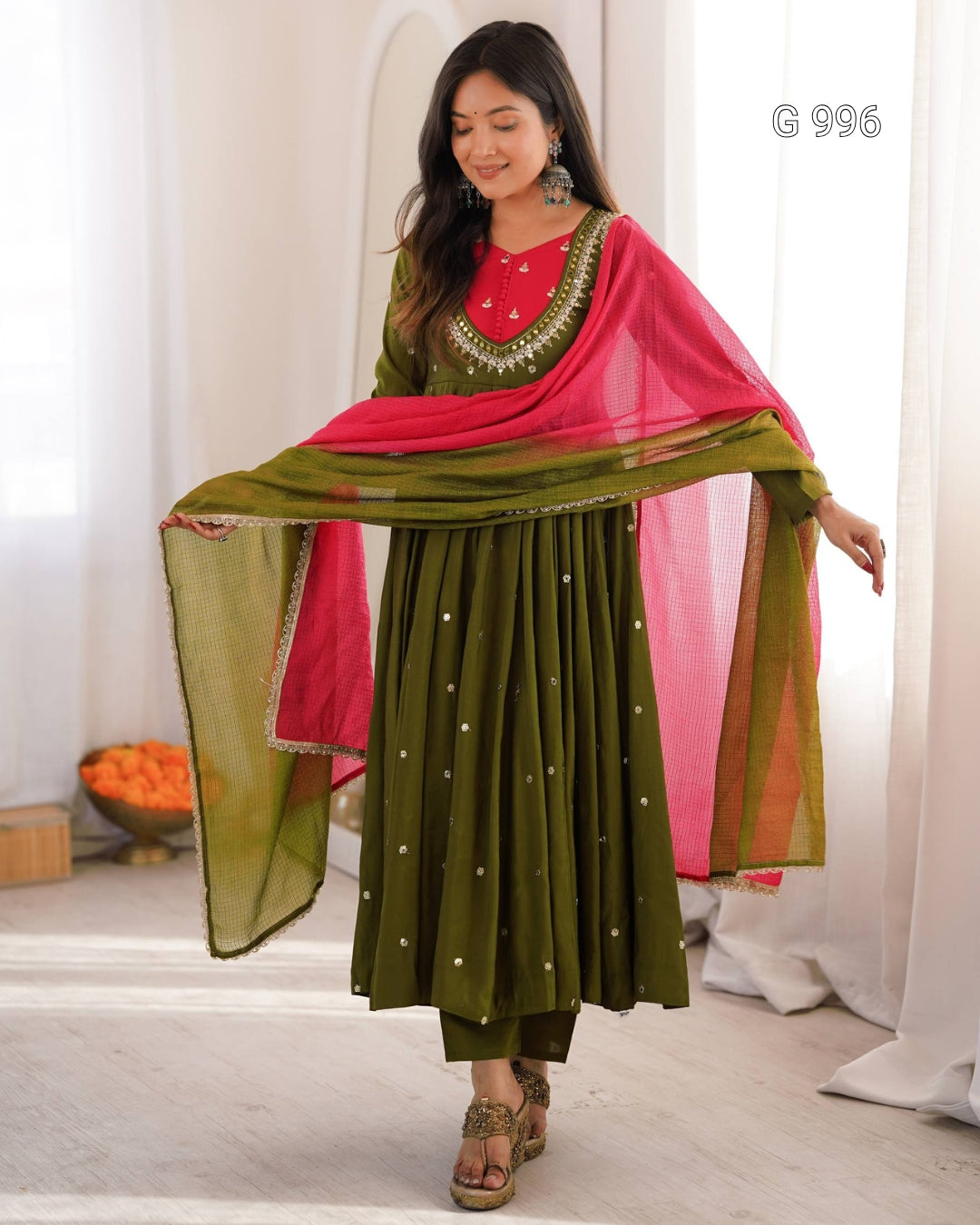 A woman modeling a traditional Indian outfit consisting of an olive green anarkali skirt with small gold dots, a matching embroidered green blouse with a bright pink embroidered neckline, and a vibrant pink dupatta draped over her shoulders.