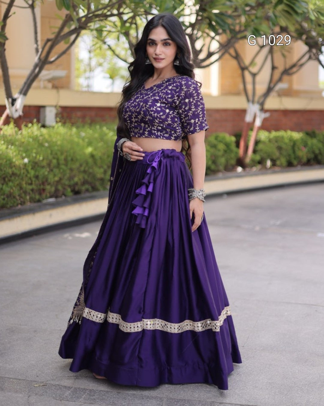 A woman wearing a purple traditional outfit with a patterned crop top and a long pleated skirt with silver trim, standing outdoors on a paved walkway.