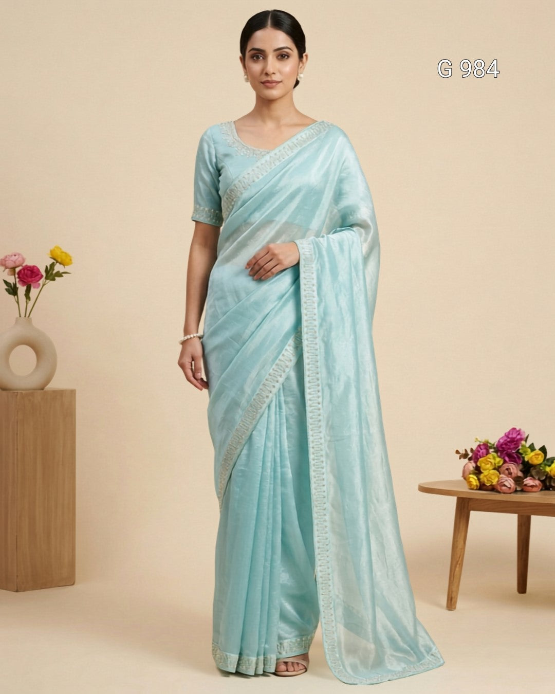A woman models a light blue-green saree with a matching short-sleeve blouse. The fabric has a subtle sheen and a decorative border.