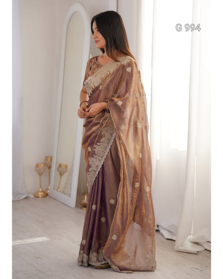 A woman wearing a metallic bronze and purple saree with gold zari embroidery, standing in a bright room with white curtains and a mirror.
