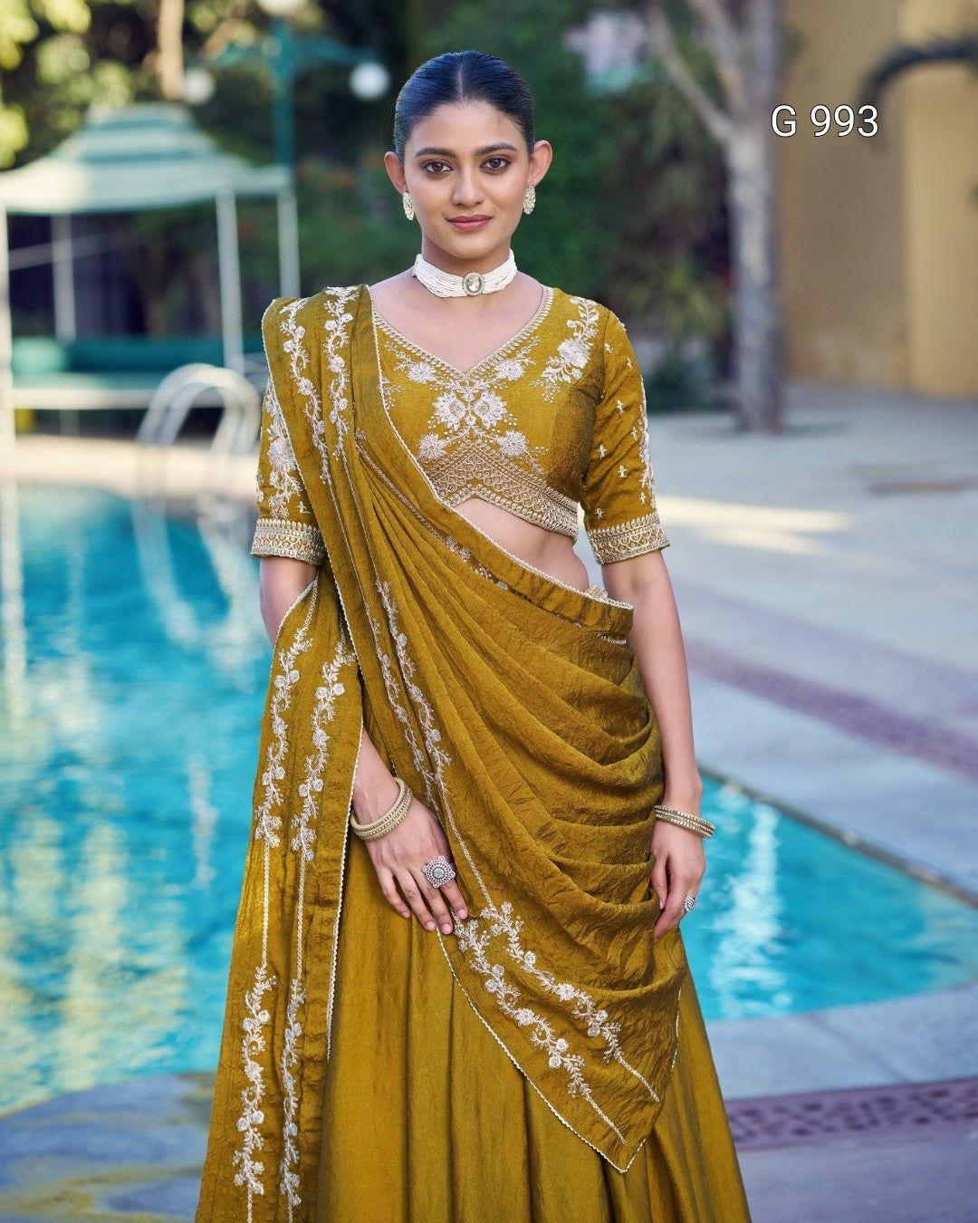 A woman stands by a swimming pool wearing an olive-yellow embroidered saree with a matching blouse. The saree has white floral embroidery along the border, and the blouse features gold and white embroidery.