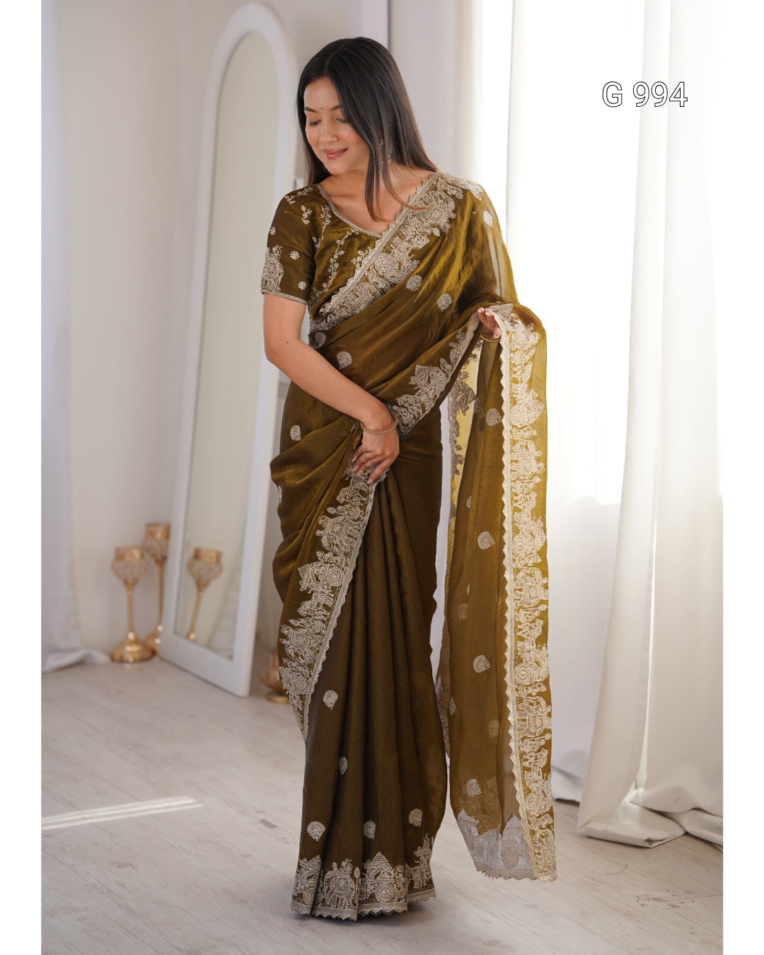 A woman models an olive green saree with intricate gold and white floral embroidery along the border and pallu, standing in a brightly lit room with white curtains.