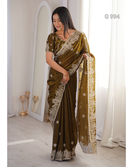 A woman models an olive green saree with intricate gold and white floral embroidery along the border and pallu, standing in a brightly lit room with white curtains.