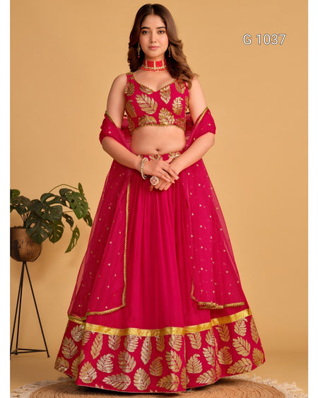 A woman modeling a bright pink lehenga choli set with gold leaf embroidery, standing against a beige background with a small potted plant on the left.
