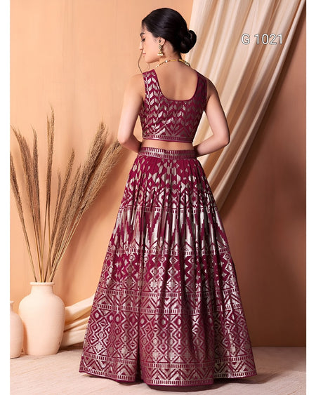 A woman modeling a deep maroon lehenga choli set with intricate silver metallic embroidery, shown from the back against a warm beige backdrop with decorative dried wheat and a vase.