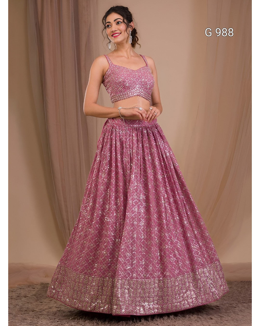 A woman models a pink embellished traditional Indian outfit consisting of a sleeveless crop top and a voluminous flared skirt, set against a neutral draped backdrop.