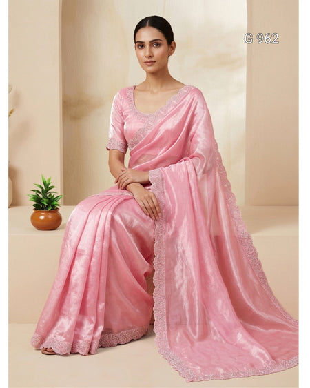 A woman seated indoors wearing a light pink saree with a matching blouse. The fabric has a subtle sheen and scalloped lace trim, with a small potted plant beside her on a beige set.