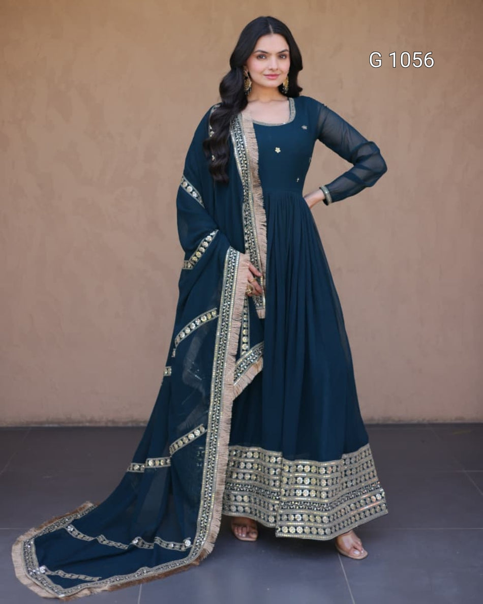 A woman models a dark teal blue anarkali gown with beige and gold embroidery, paired with a matching dupatta, set against a plain beige wall.