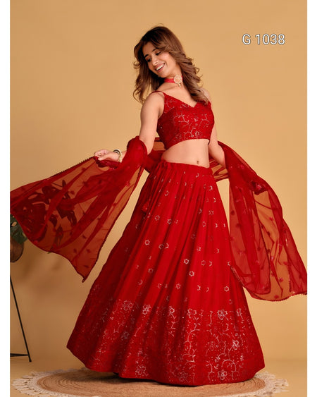 A woman modeling a red embroidered Indian ethnic wear set consisting of a flared lehenga skirt, a matching crop top, and a sheer dupatta, set against a solid beige background.