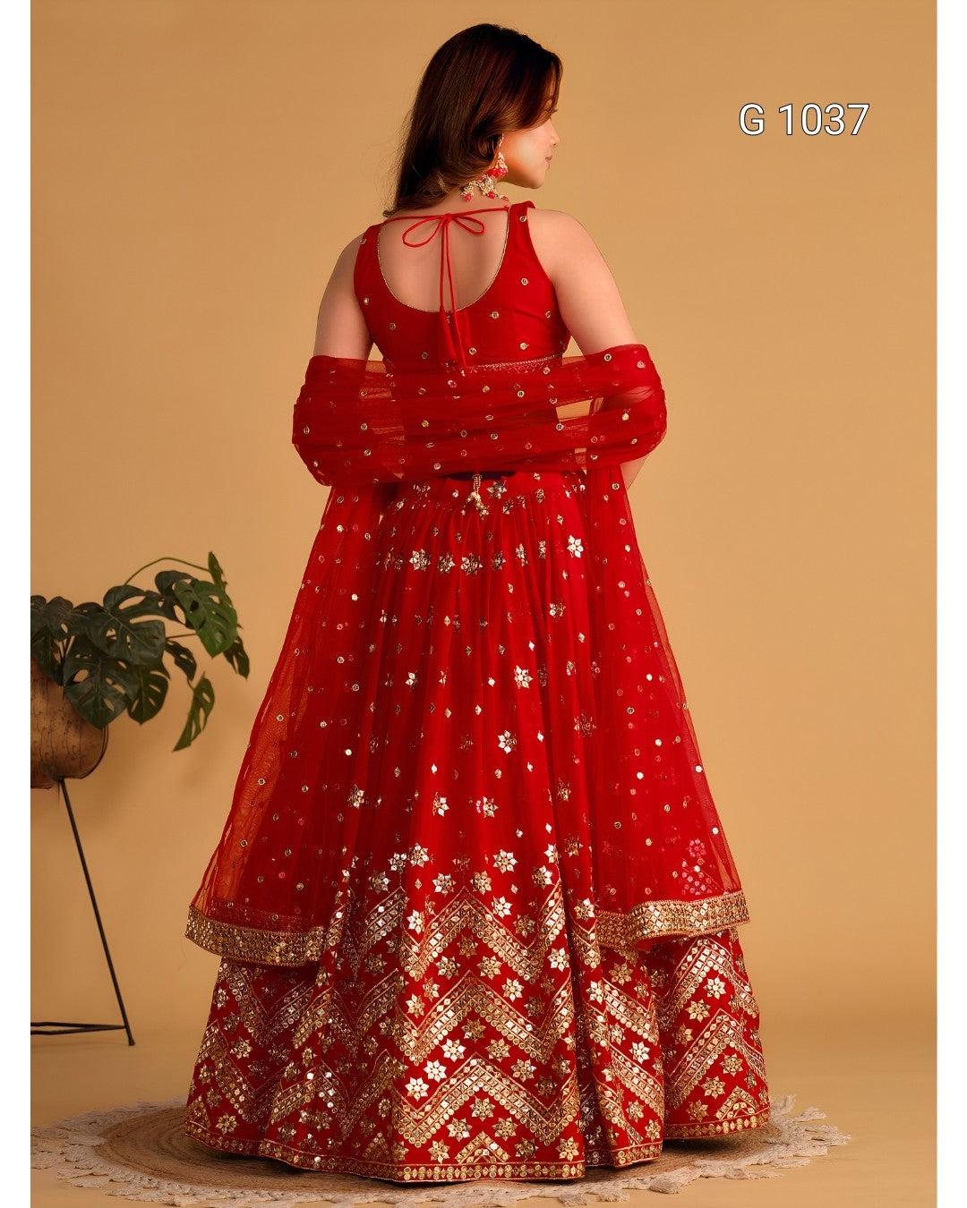 A woman wearing a red traditional Indian dress with gold embroidery, shown from the back against a beige studio background. The dress has sheer sleeves, a tie at the back neckline, and ornate gold patterns concentrated around the lower skirt.