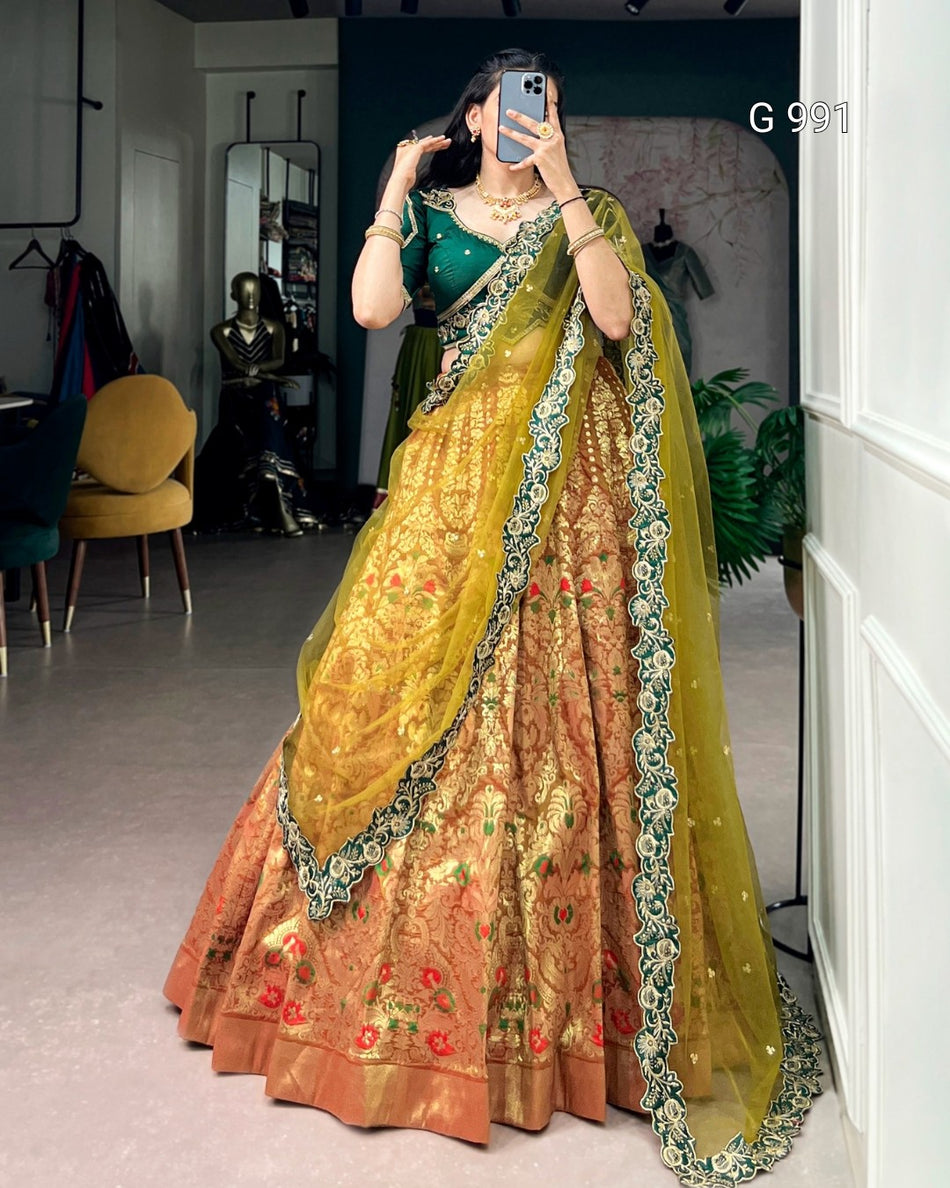 A woman takes a mirror selfie wearing an ornate traditional Indian lehenga choli outfit. The outfit features a deep green sleeveless blouse, a heavily embroidered peach and gold flared skirt, and a sheer yellow-green dupatta with green floral borders.