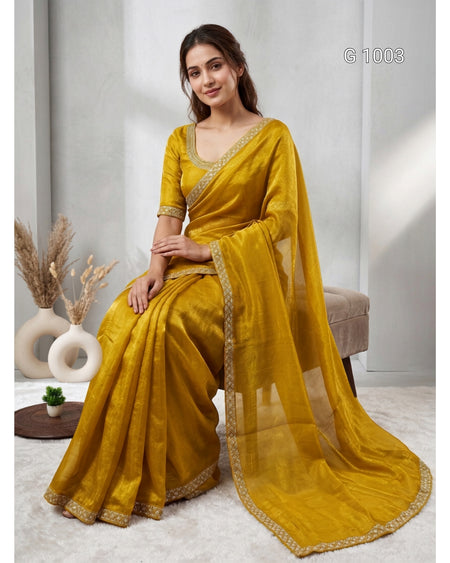 A woman models a mustard yellow saree with a matching blouse and dupatta, featuring gold embroidered borders, seated in a styled indoor setting.