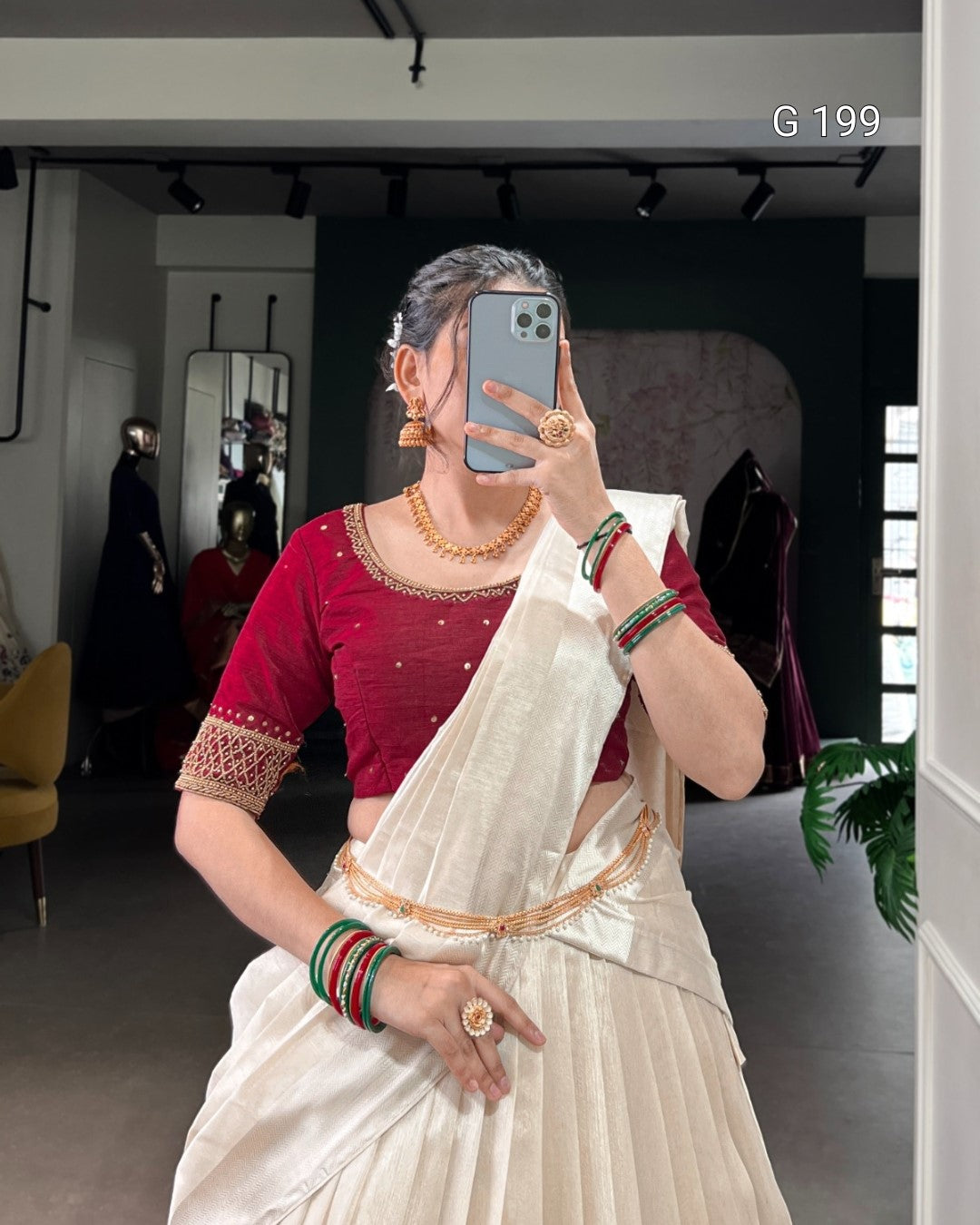 Woman taking a mirror selfie wearing a red blouse and white saree with gold border.