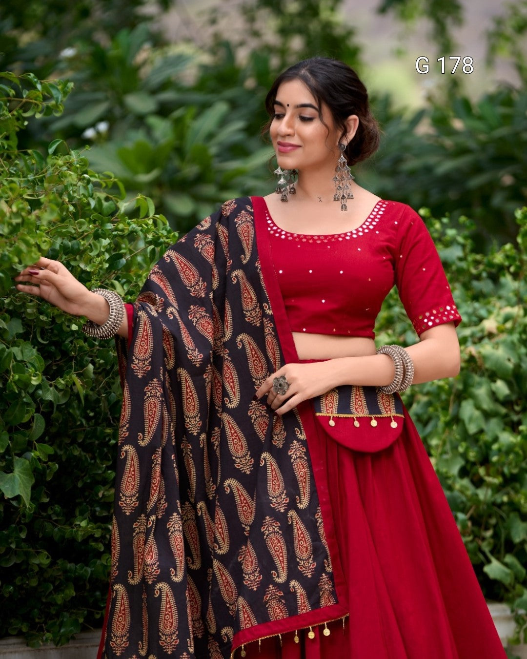 Woman wearing a red traditional outfit with a black and orange patterned shawl against a green leafy background.