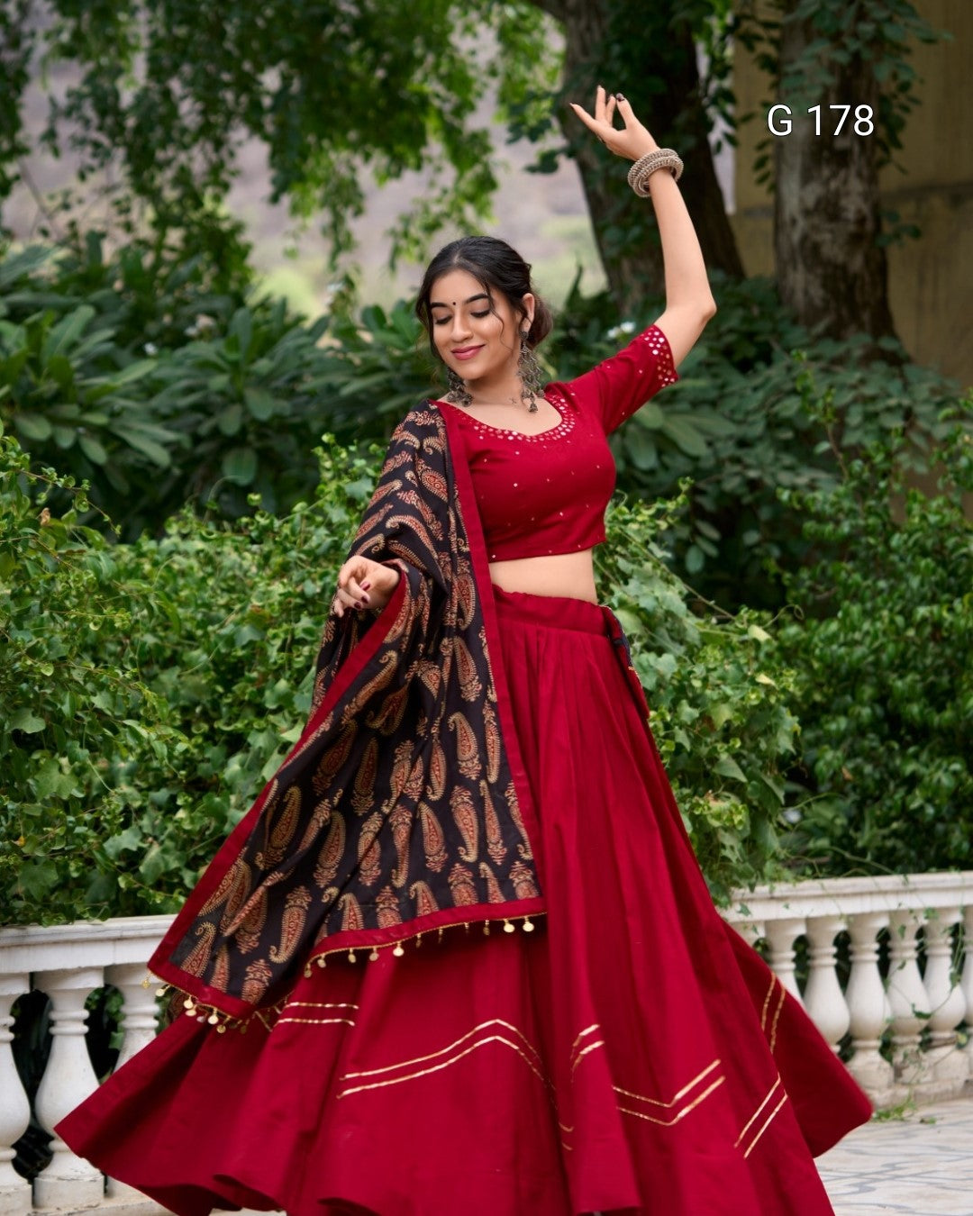 Woman in a red traditional outfit with a patterned dupatta, standing outdoors with greenery in the background.